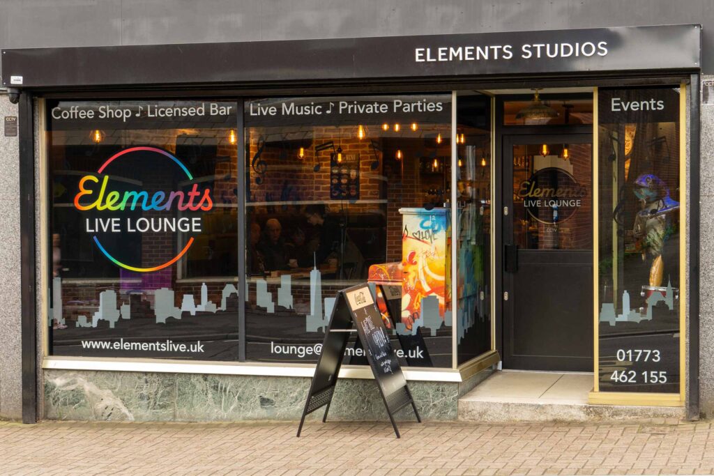 Photograph of Elements Live Lounge shop front showing entrance and windows decorated with large logo, musical notes and building skyline vinyl graphics.